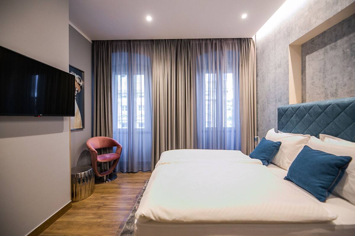 Hotelkamer met een groot bed, een flatscreen televisie aan de wand, een rode fauteuil en uitzicht op de stad door grote ramen.