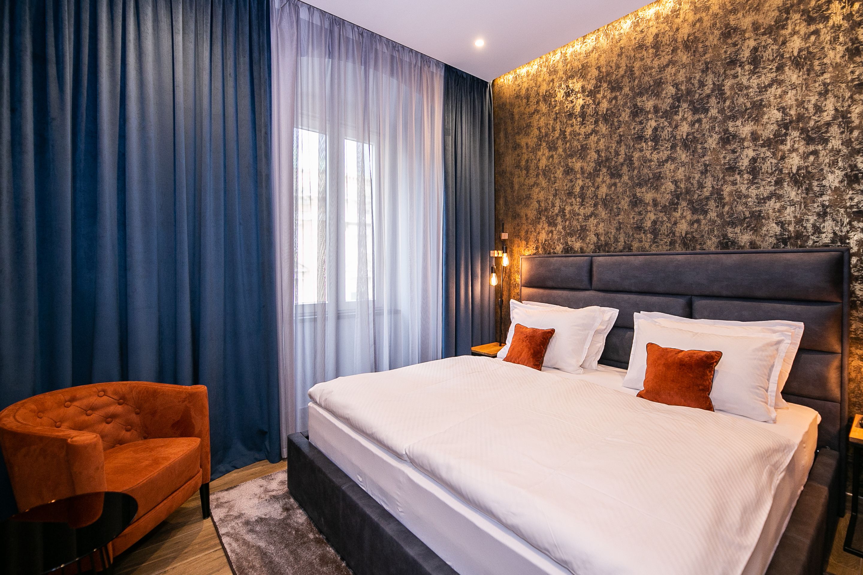 Une chambre d’hôtel moderne avec un grand lit, un fauteuil orange à motifs et un mur à texture.
