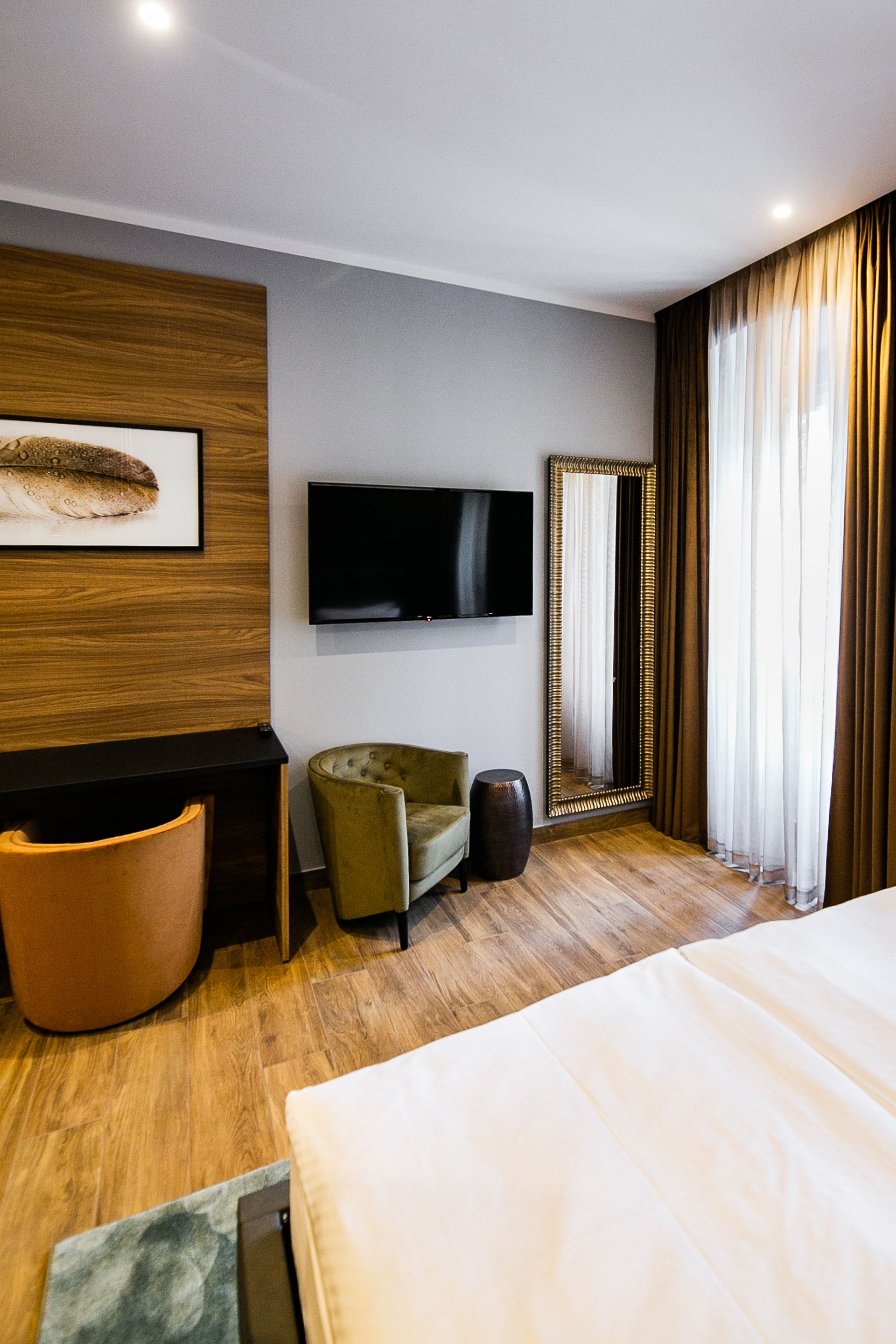 Een hotelkamer met een bed, een aan de muur bevestigde televisie, een grote spiegel, een groene fauteuil en een houten vloer.