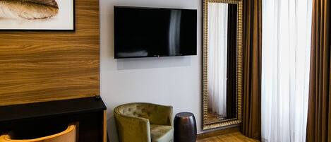 A hotel room with a bed, a television mounted on the wall, a large mirror, a green armchair, and a wooden floor.