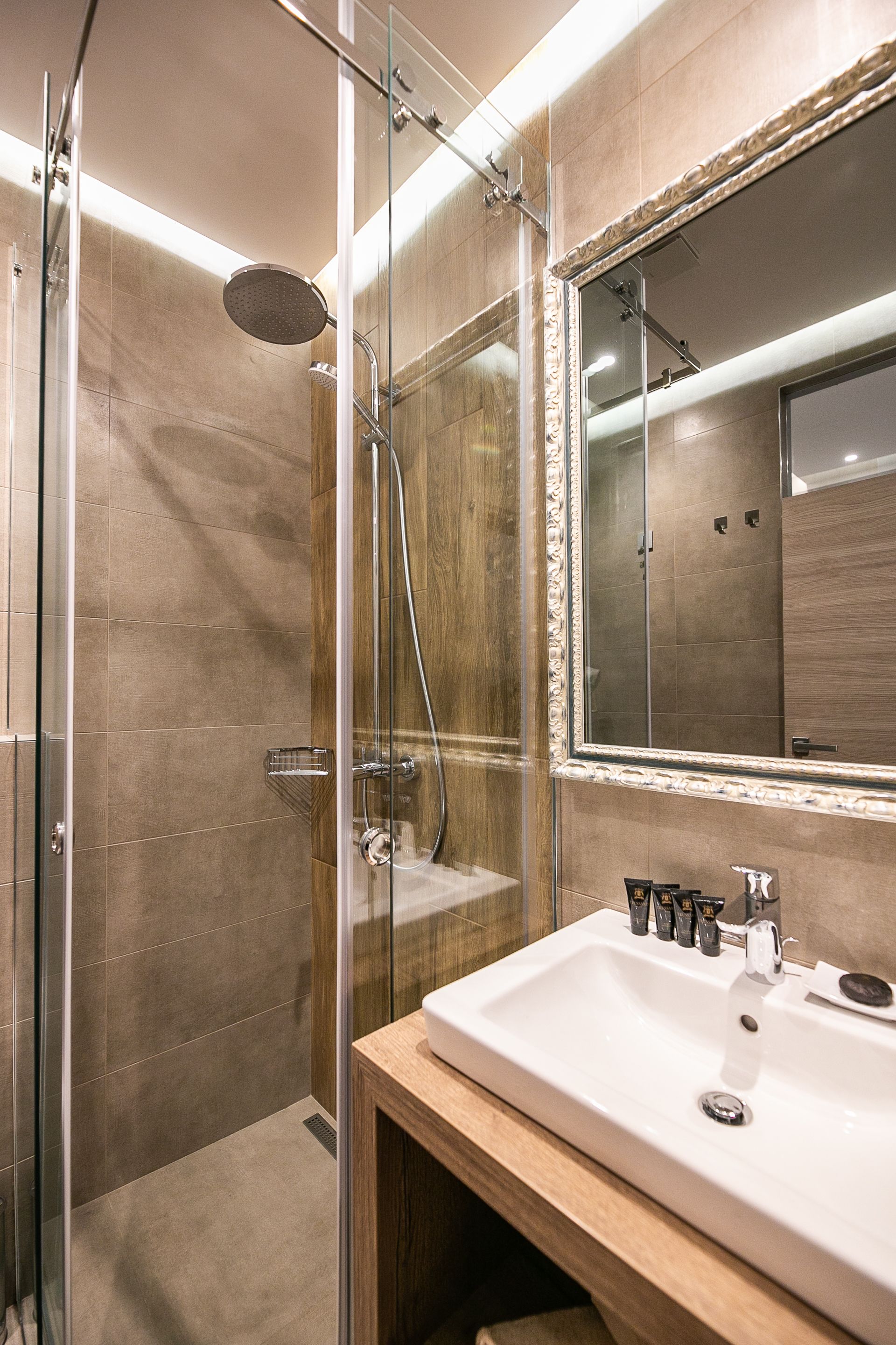 Et moderne badeværelse med et glasafskærmet brusebad, et stort spejl og en hvid vask med toiletartikler.