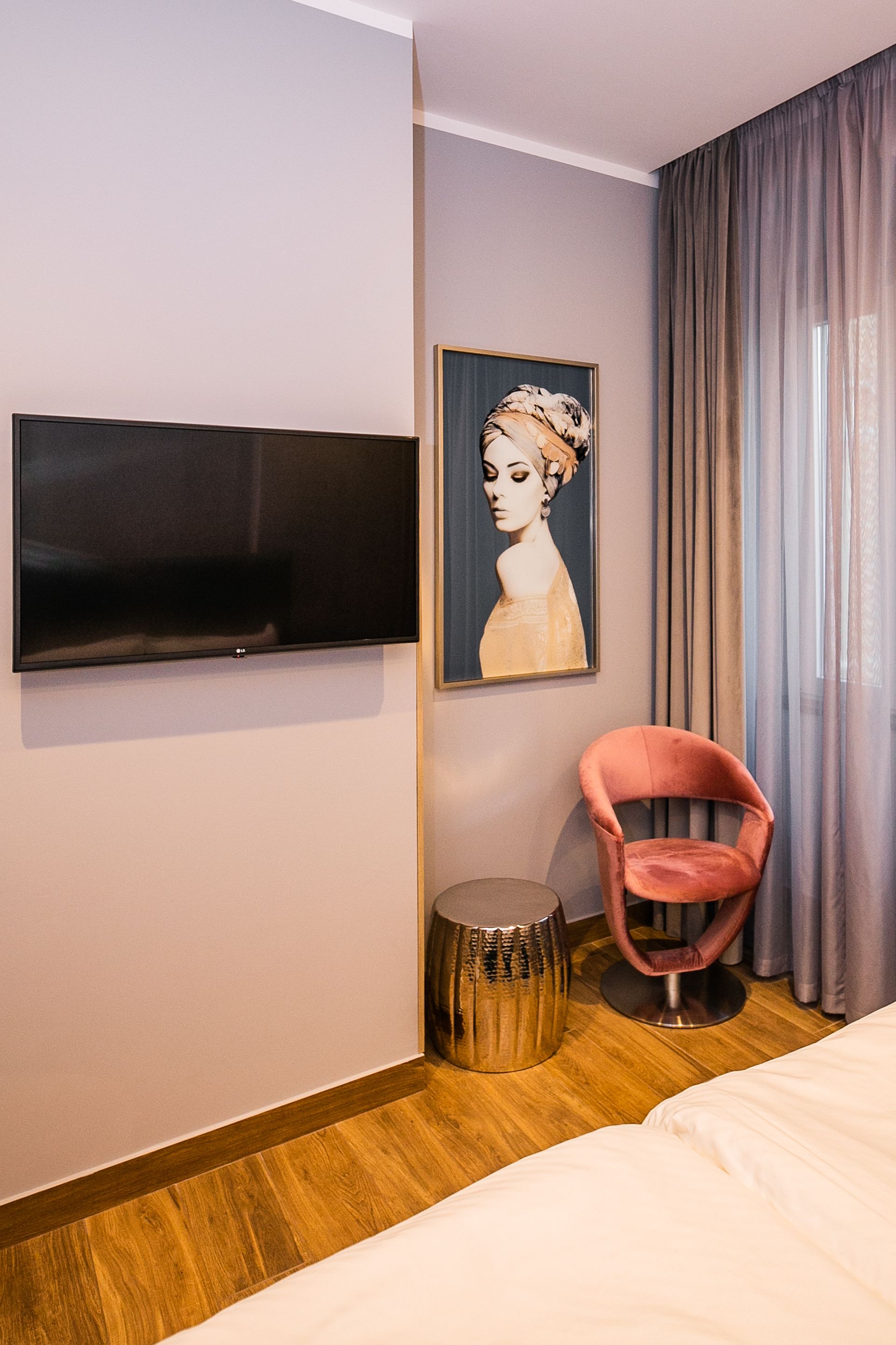 Ein Hotelzimmer mit einem an der Wand befestigten Flachbildfernseher, einem gerahmten Porträt, einem rosa Sessel, einem kleinen runden Beistelltisch und einem Bett mit weißer Bettwäsche.