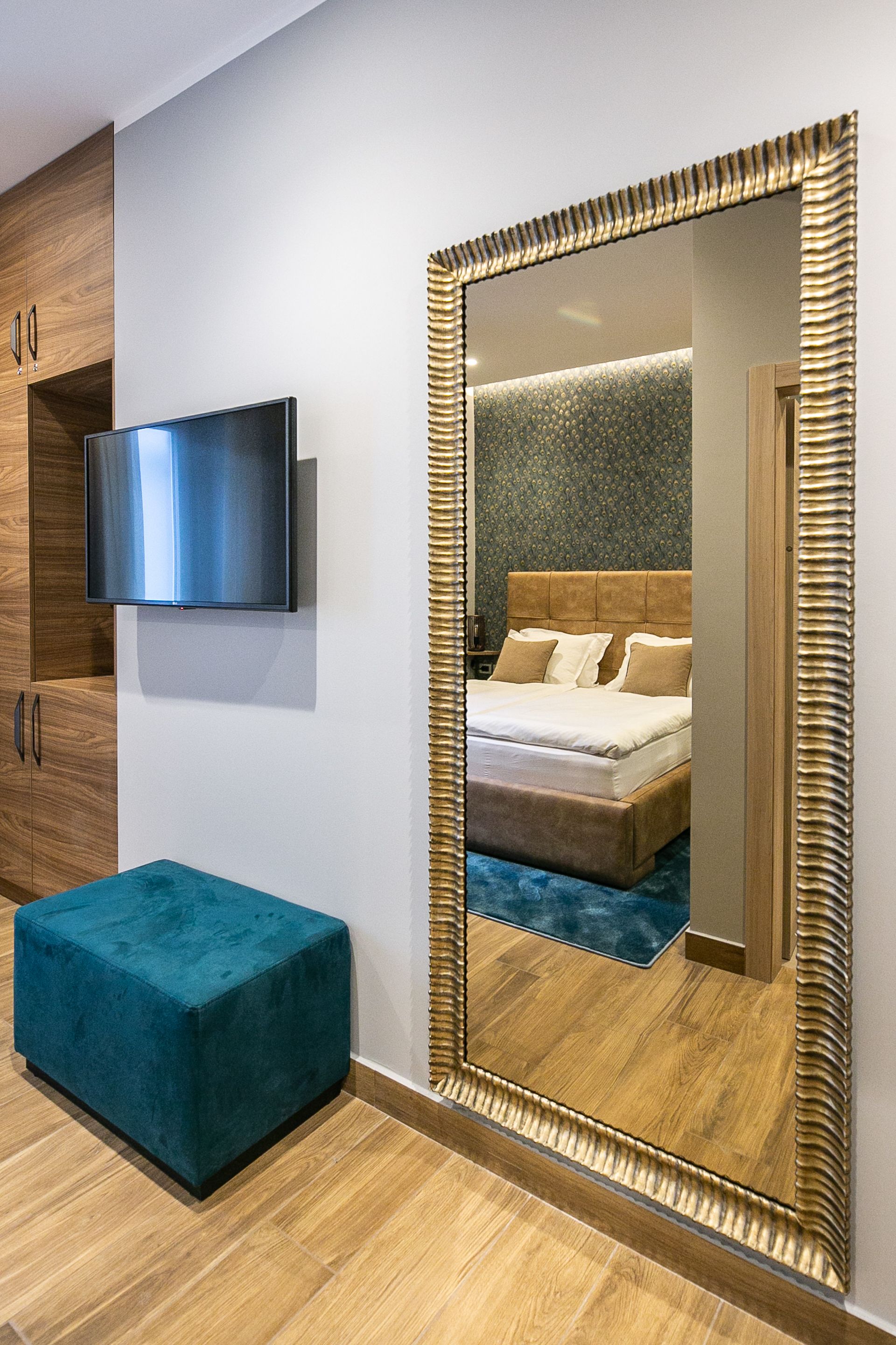 A modern bedroom with a large mirror, a flat-screen TV, wooden cabinets, and a teal ottoman.
