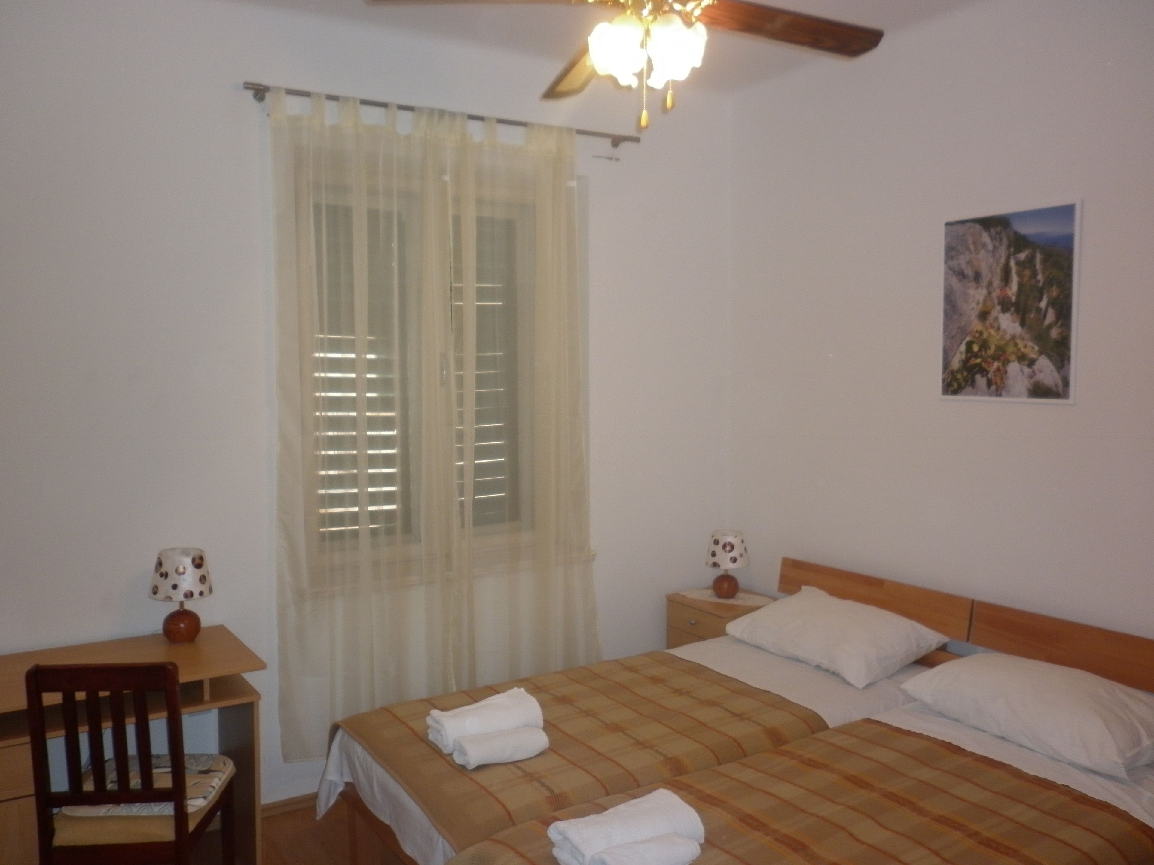 Un dormitorio con cama, escritorio, silla, mesita de noche, lámpara y un cuadro en la pared.