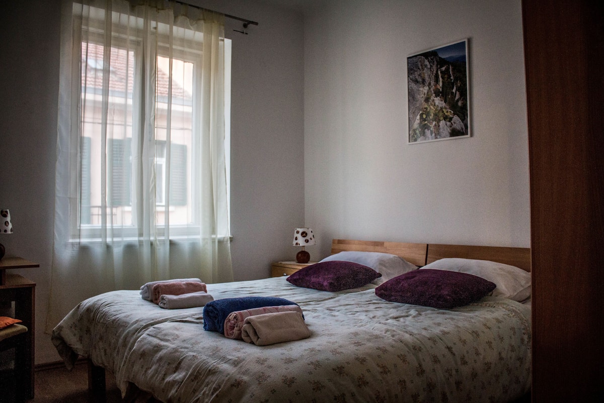 A bedroom with two beds, a window with sheer curtains, a nightstand with a lamp, and a painting on the wall.