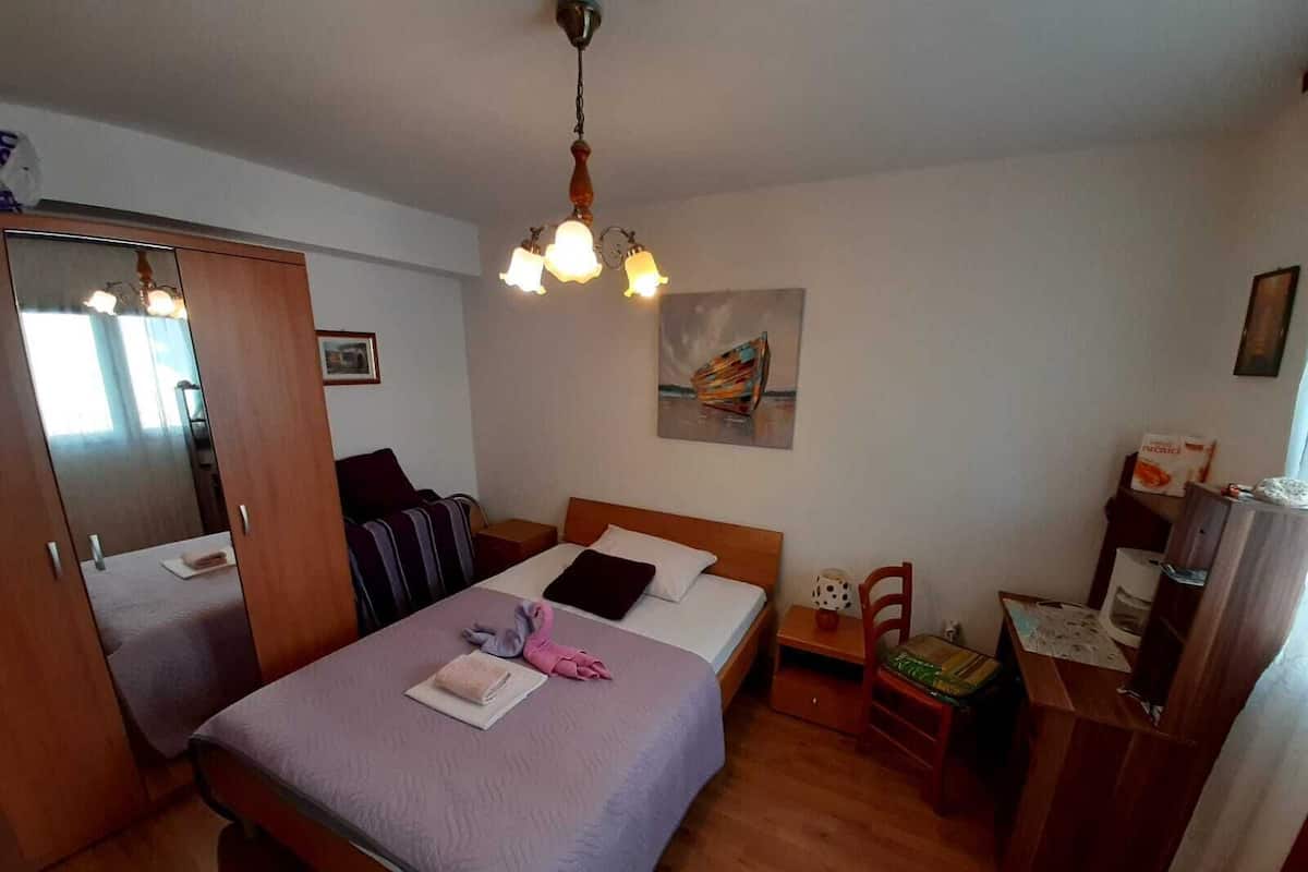 A bedroom with a bed, a wooden wardrobe, a nightstand, a chair, a desk, a hanging light fixture, and a painting on the wall.