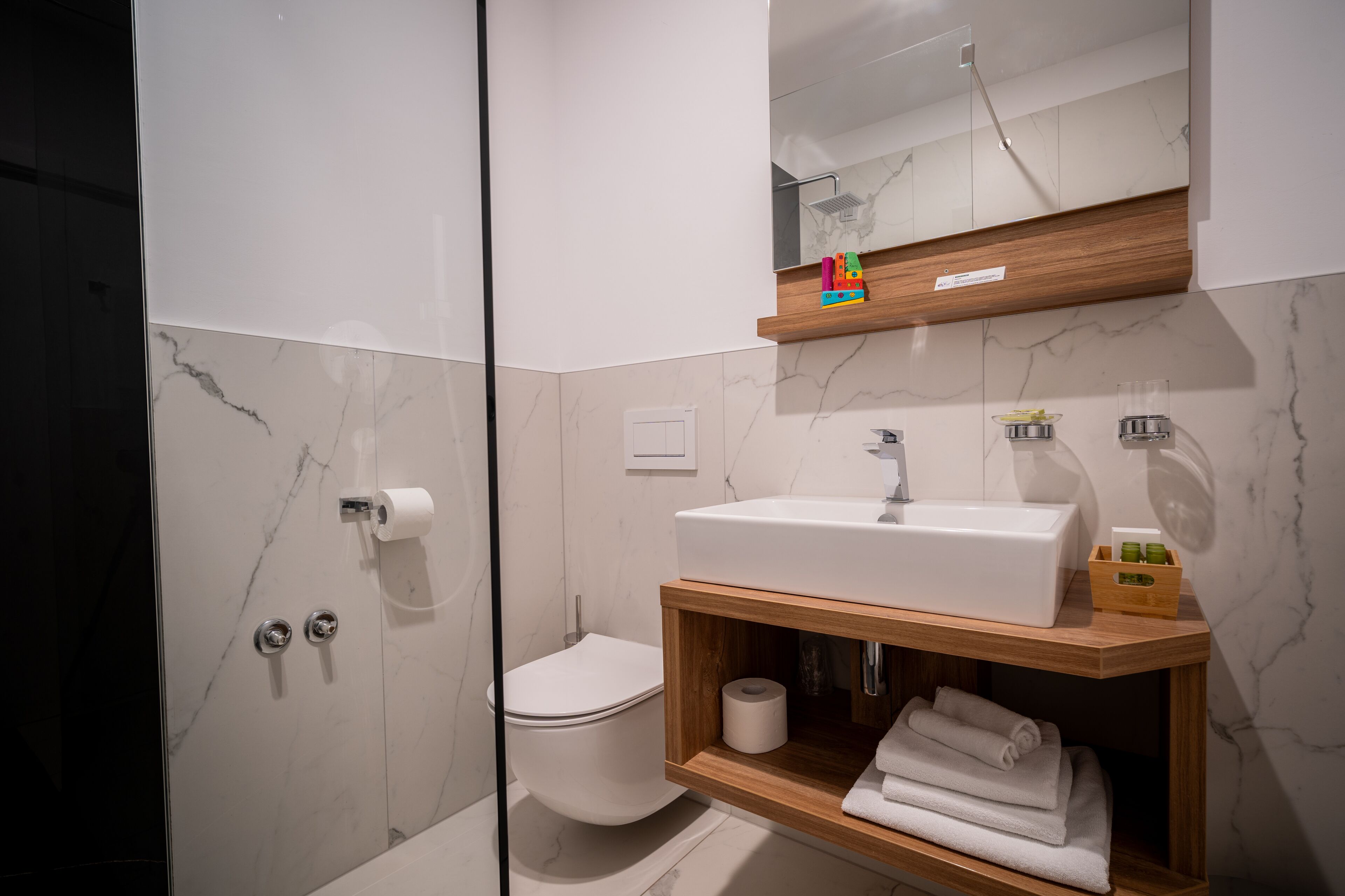 Une salle de bain moderne avec une douche à l’italienne en verre, des toilettes blanches, un meuble-lavabo en bois et un miroir au-dessus du meuble-lavabo.