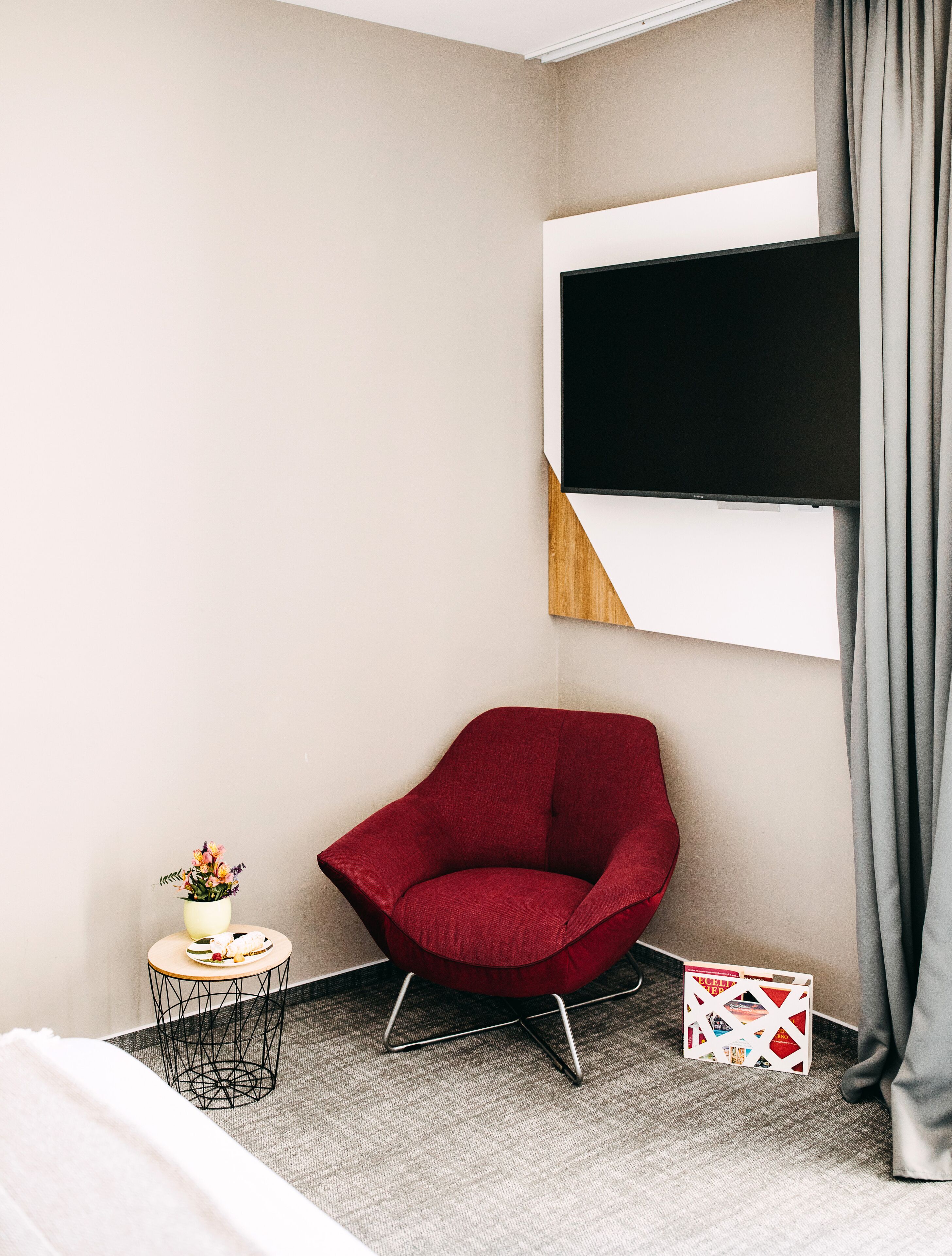 A modern hotel room with a red armchair, a small round table with a vase of flowers, and a flat-screen TV mounted on the wall.
