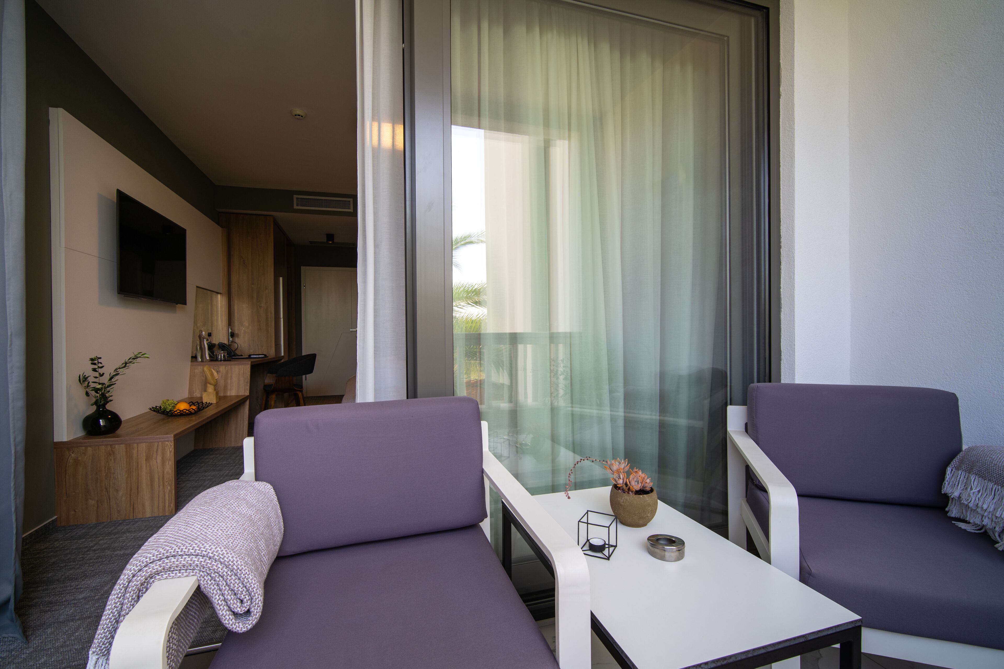 A modern room with two purple sofas, a small white table, and a glass door leading to a balcony.