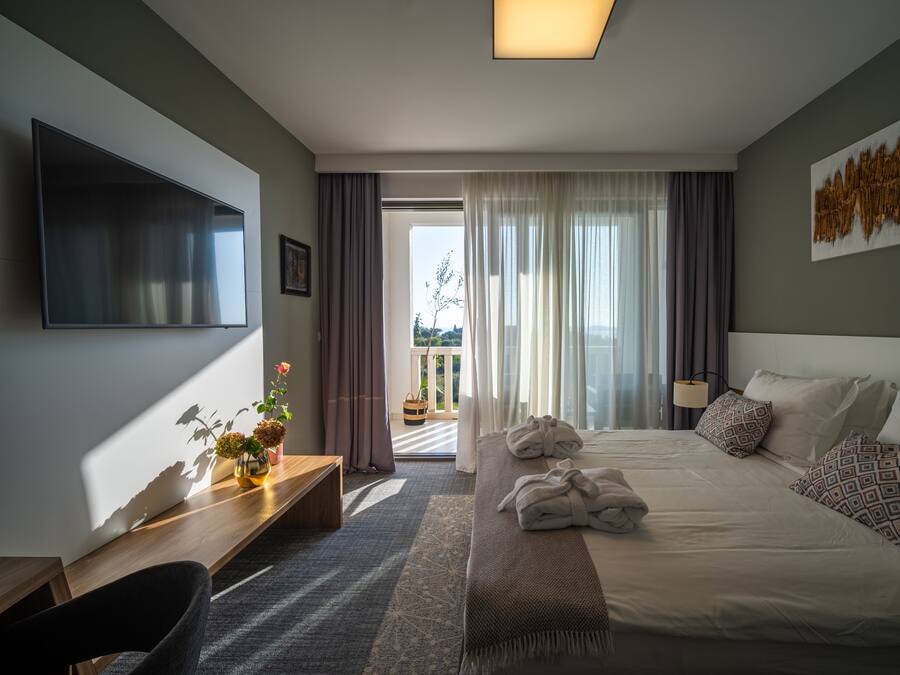 A modern hotel room with a large bed, a desk, a flat-screen TV, and a balcony with curtains.