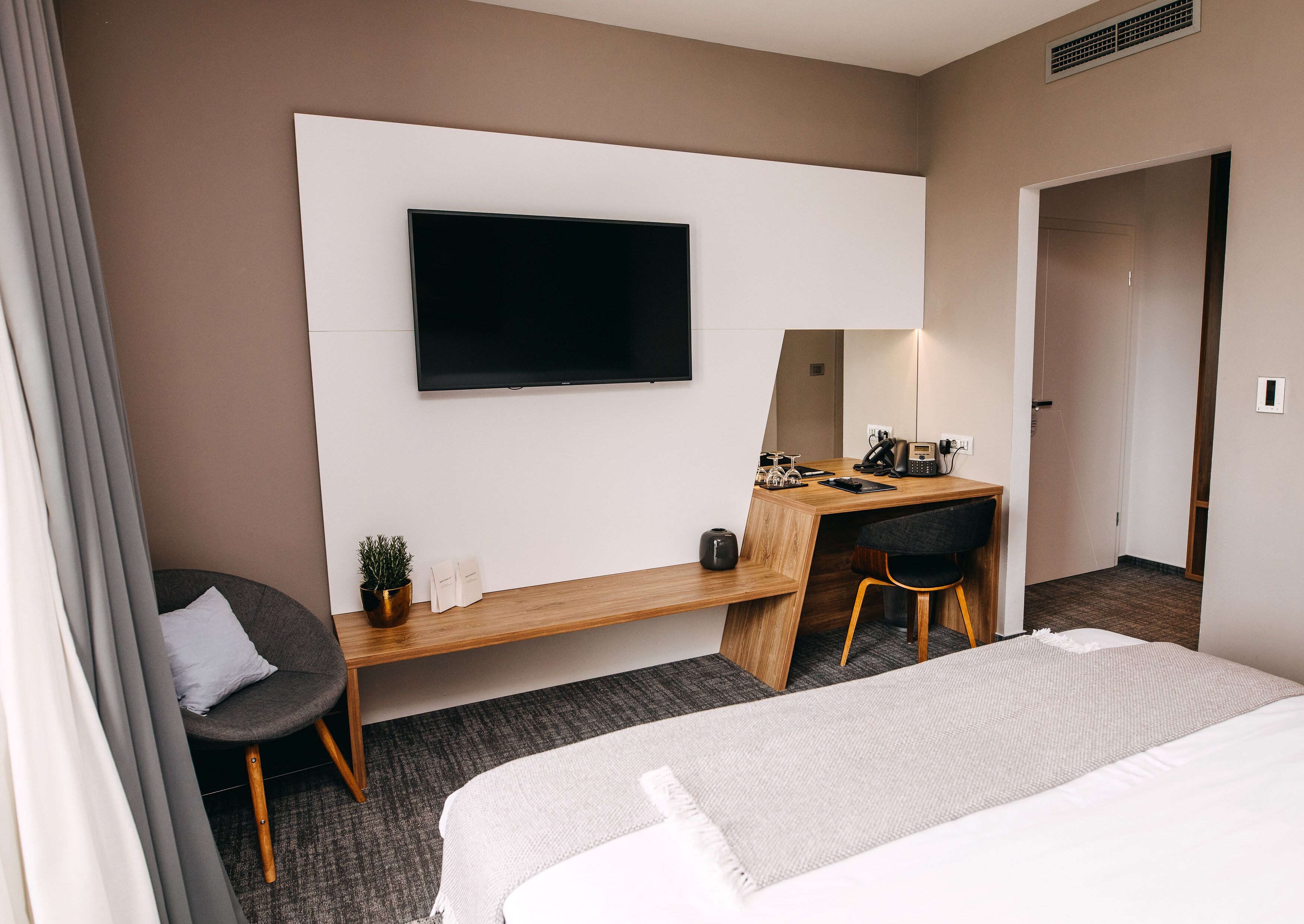 A hotel room with a bed, a desk with a chair, a television, and a small potted plant.