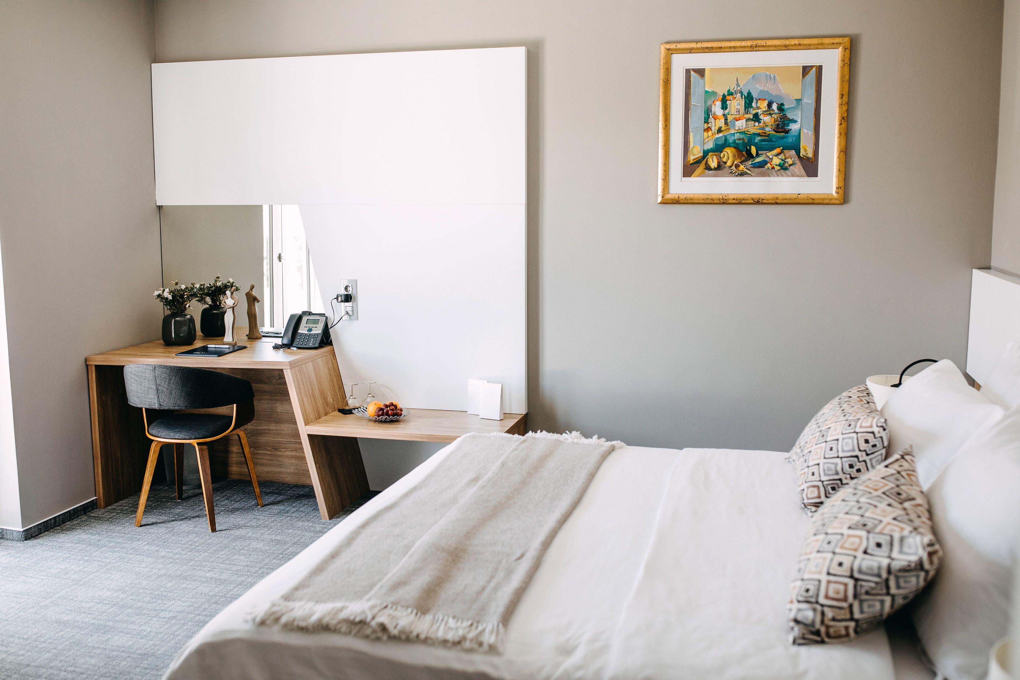 A bedroom with a bed, a desk, a chair, a mirror, and a painting on the wall.