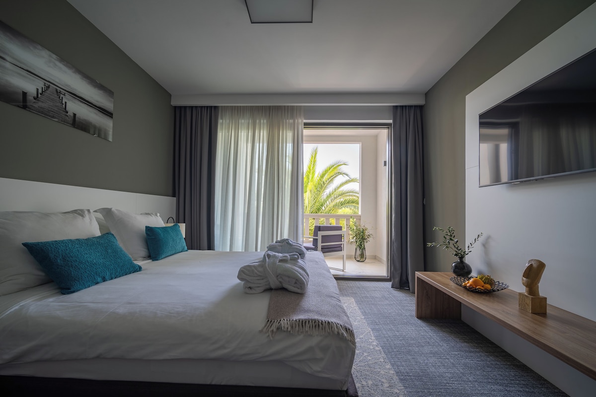 A modern hotel room with a large bed, a TV, and a balcony with a view of palm trees.