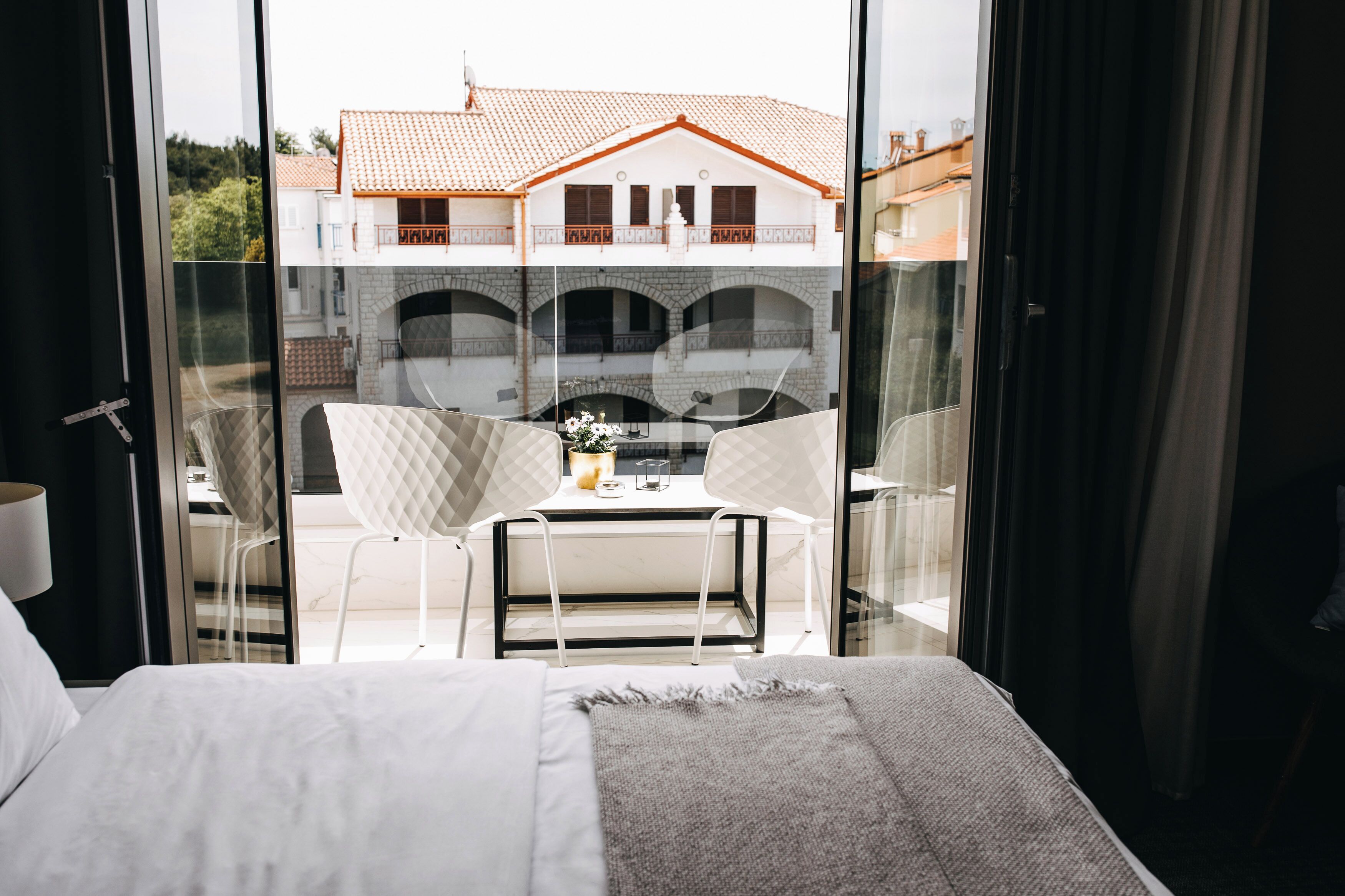 Quarto de hotel com uma cama, uma mesinha com cadeiras e vista para um edifício com telhado de azulejos através de uma porta de vidro.