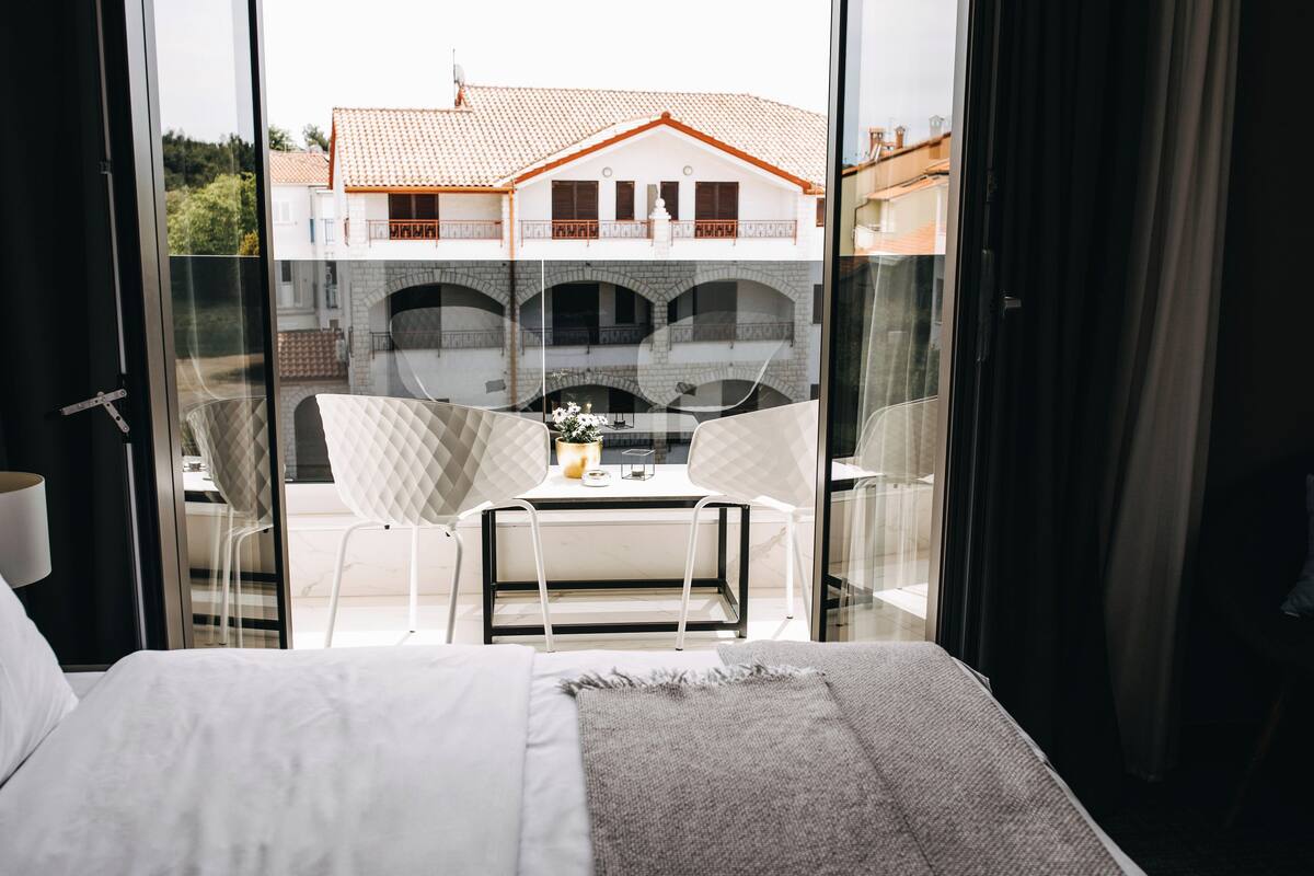 A hotel room with a bed, a small table with chairs, and a view of a building with a tiled roof through a glass door.