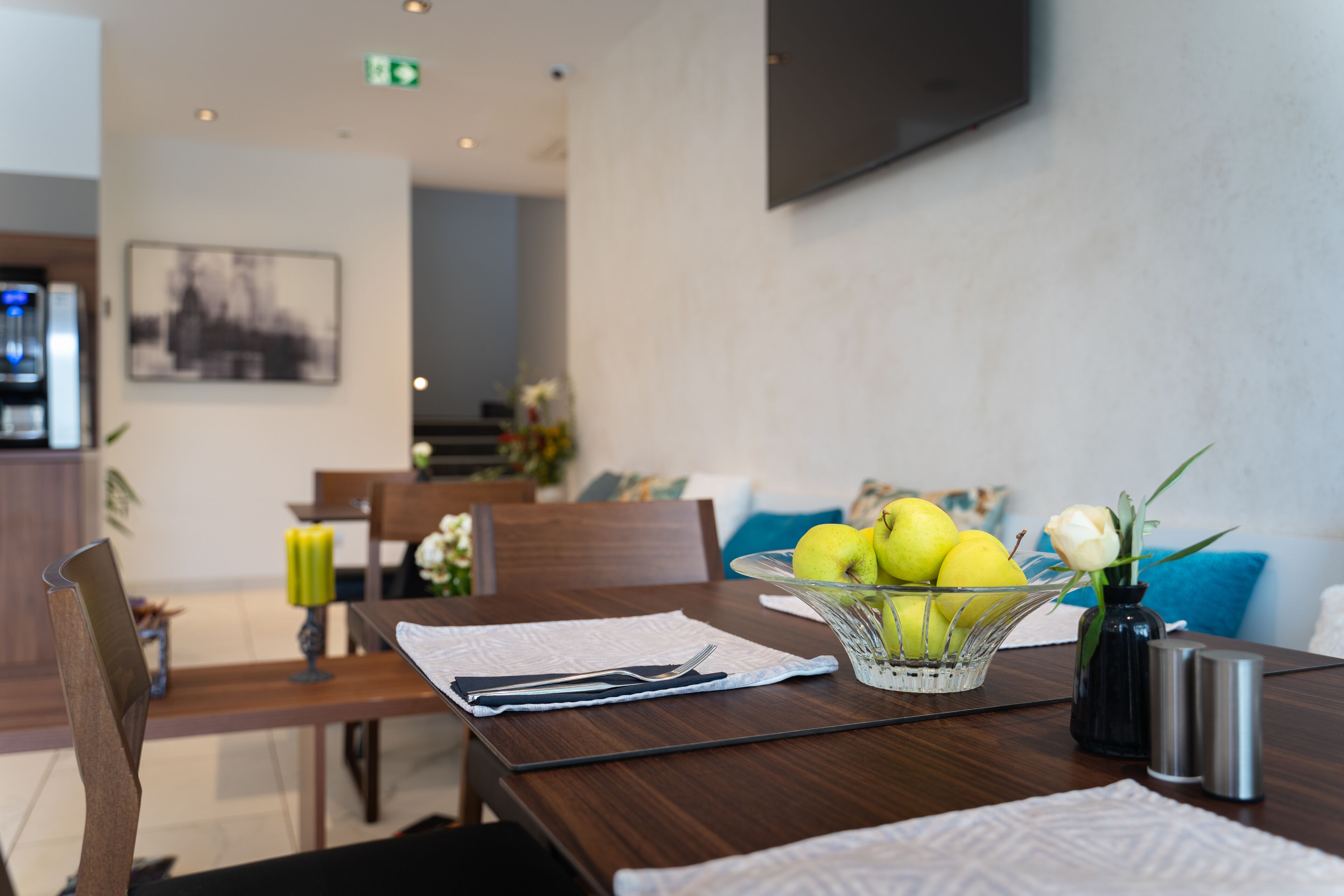 A dining area with a table set for two, a bowl of fruit, a vase with a flower, and a television mounted on the wall.