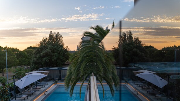 Una piscina con lettini e ombrelloni, circondata da alberi e un cielo al tramonto.