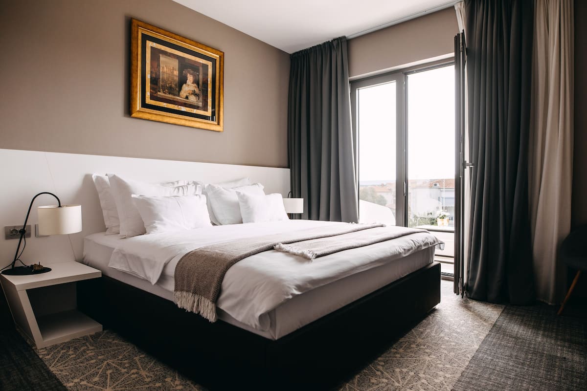 A hotel room with a large bed, a nightstand, a lamp, a framed picture on the wall, and a view of the city through the window.
