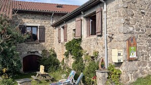 A stone building with a rustic appearance, featuring a wooden door and shuttered windows. There is a wooden bench and a lounge chair in the foreground, and a sign indicating a location or point of interest. - Feng Shui Cottage in Marlhes' Nature (Marlhes)