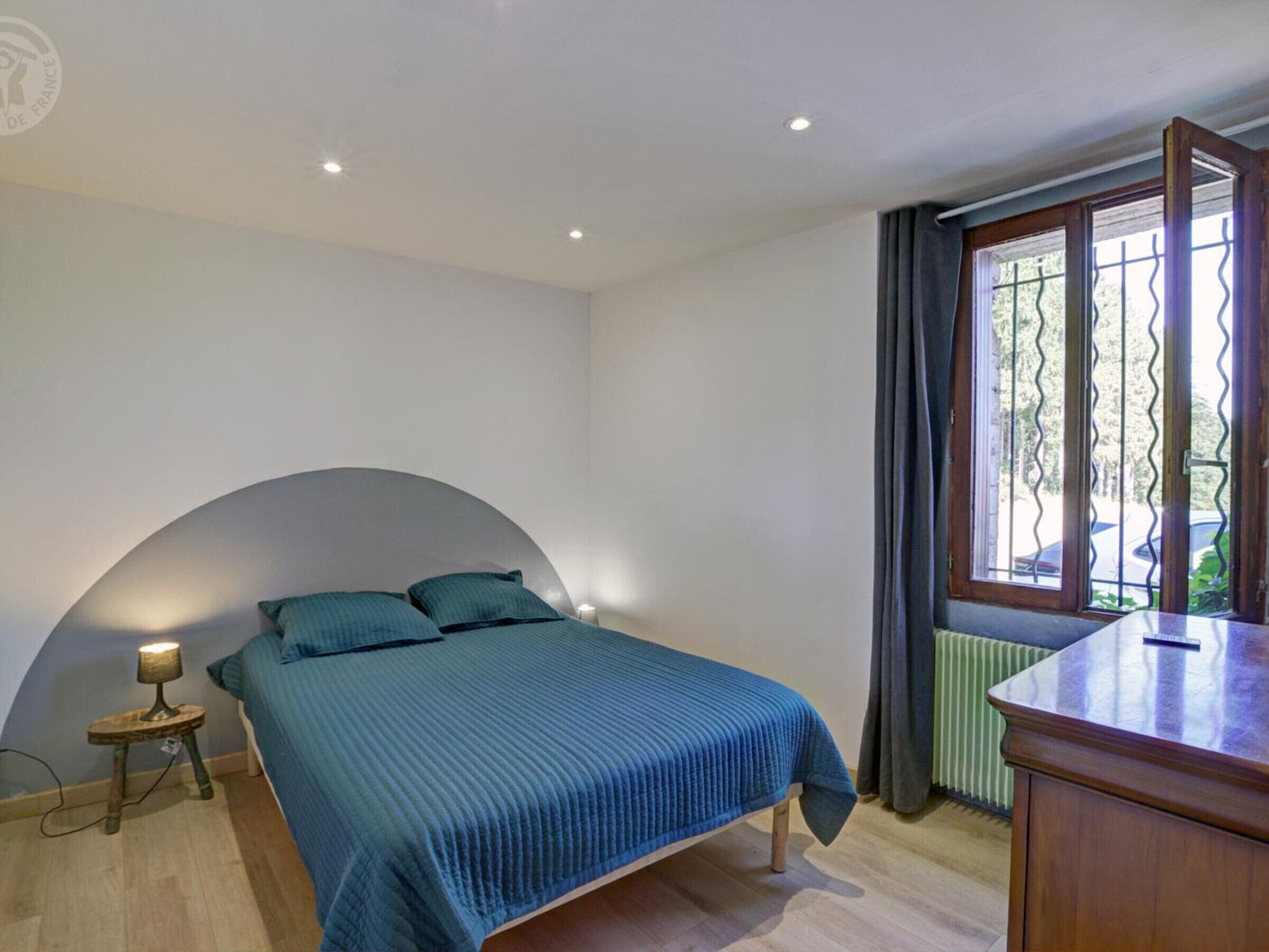 A bedroom with a curved headboard, a bed with a blue bedspread, a bedside table with a lamp, a window with curtains, and a radiator.