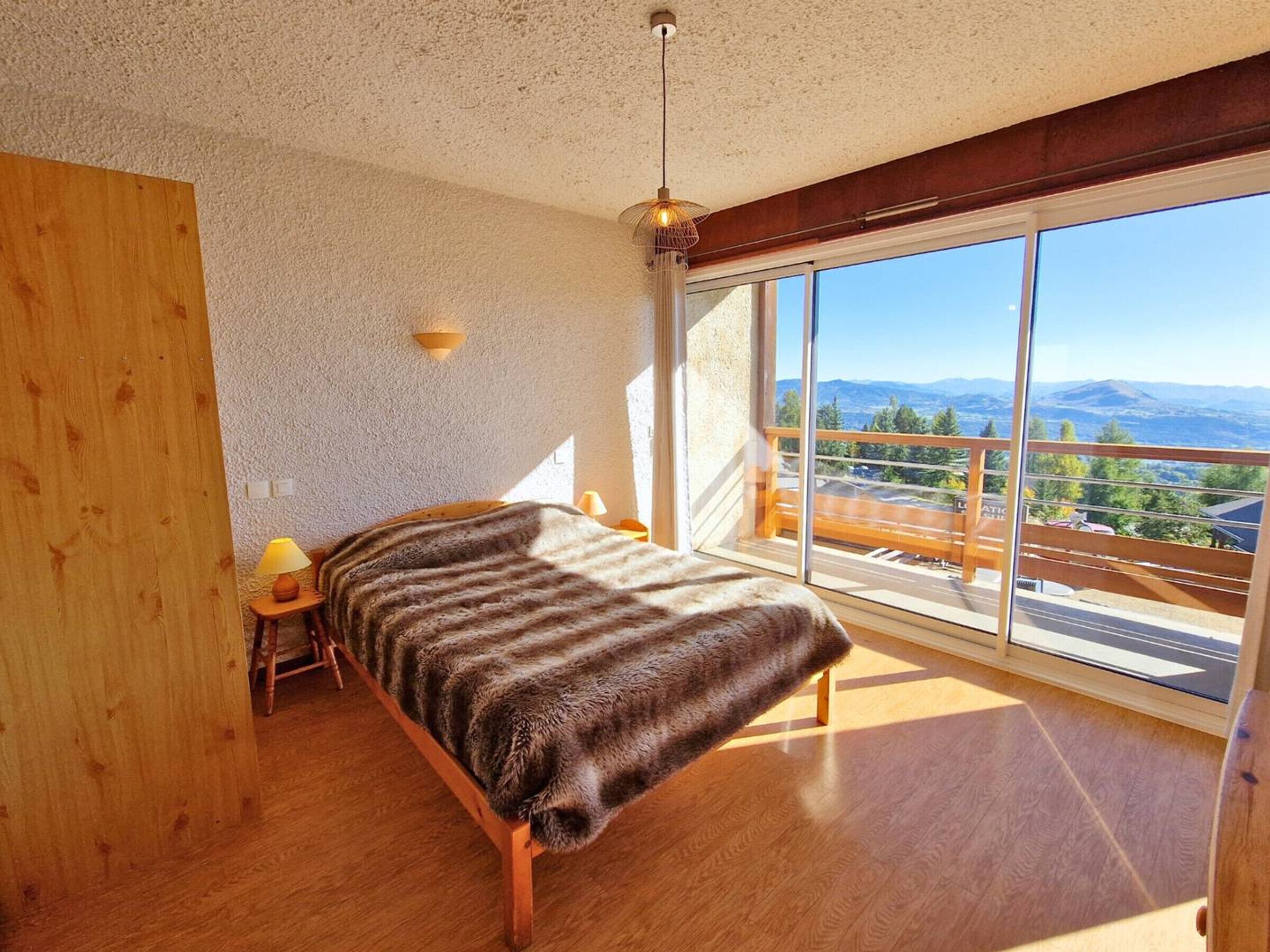 Un dormitorio con un ventanal grande, una cama de madera, una mesita de noche y una lámpara.