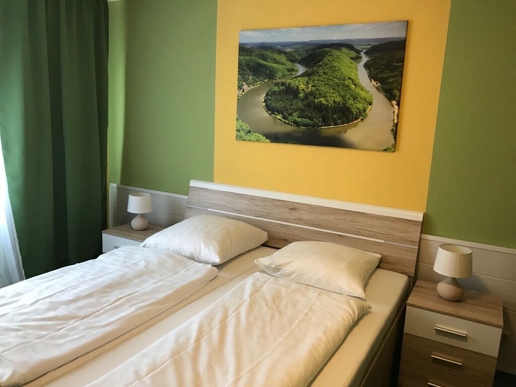 Un lit bien fait, avec du linge de lit blanc, une table de chevet avec une lampe et un tableau représentant un paysage fluvial accroché au mur.