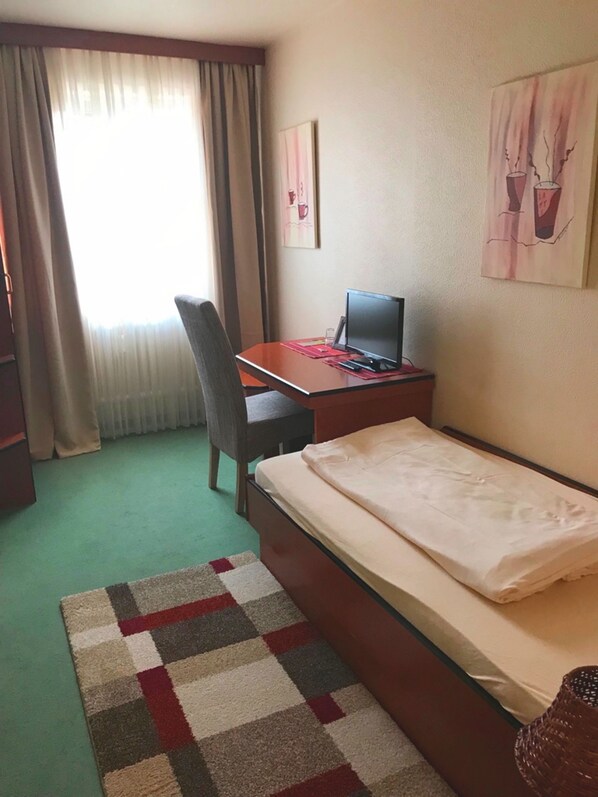 A small hotel room with a single bed, a desk with a laptop, a chair, and a window with curtains.