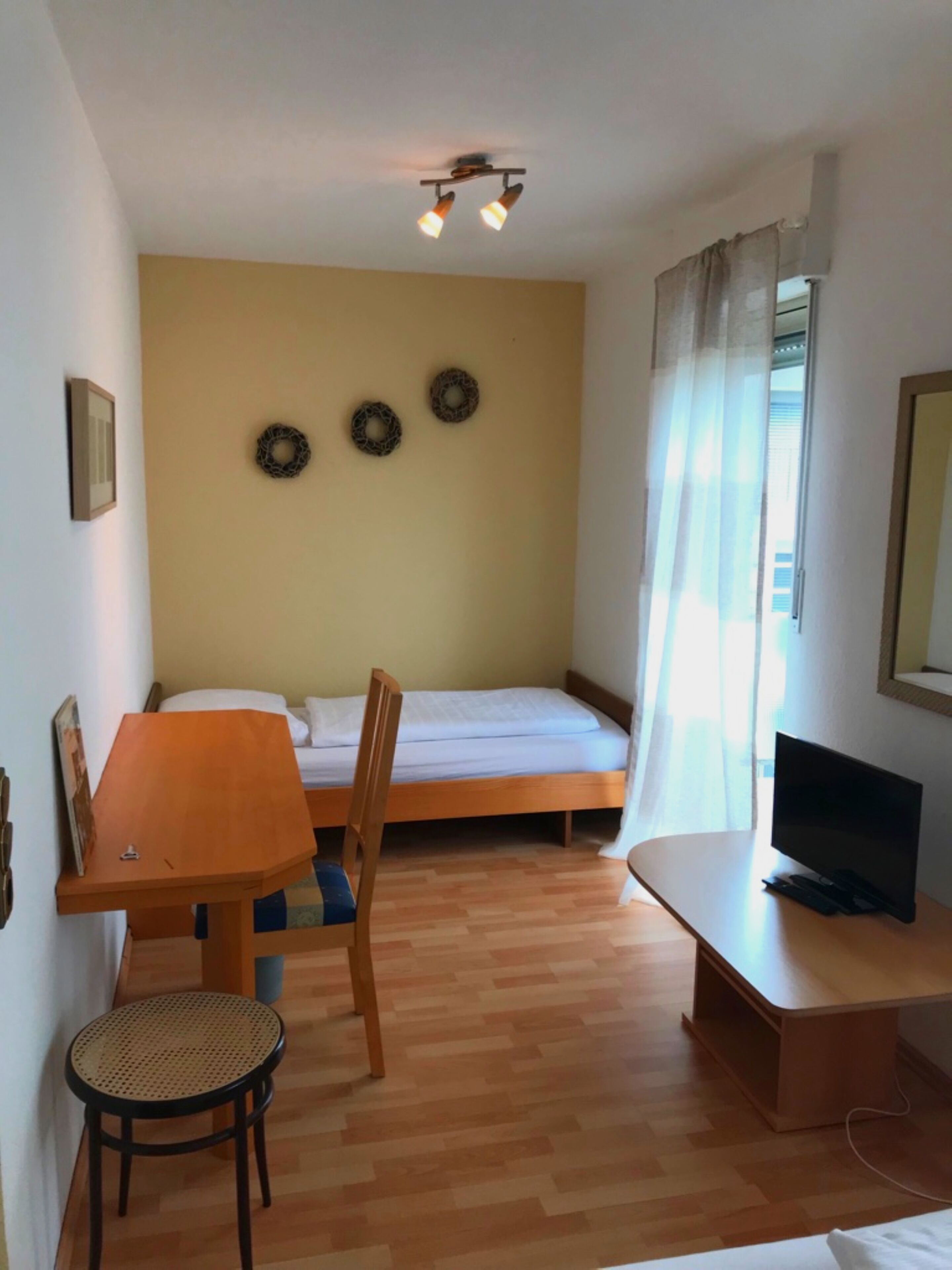 A small, well-lit room with a bed, a desk with a chair, a television, and a window with curtains.
