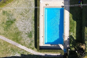 An aerial view of a rectangular swimming pool with a tiled deck surrounding it, a paved pathway, and a small grassy area.