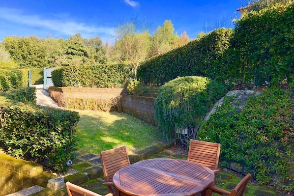 A garden with a wooden table and chairs, surrounded by lush greenery and a stone wall.