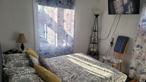 A bedroom with a bed, a chair, a floor lamp, a TV, and a window with floral curtains.