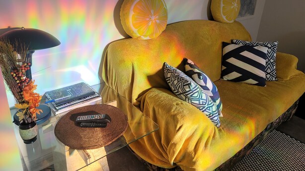 A yellow sofa with patterned pillows, a remote control on a woven coaster, a vase with dried flowers, and a bookshelf with books and a lamp.