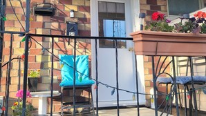 A brick house with a white door, a small porch with a blue chair, a table, and potted plants.