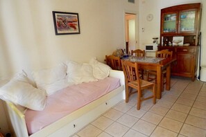 A small room with a sofa, a dining table with chairs, a kitchen area with cabinets, and a clock on the wall.