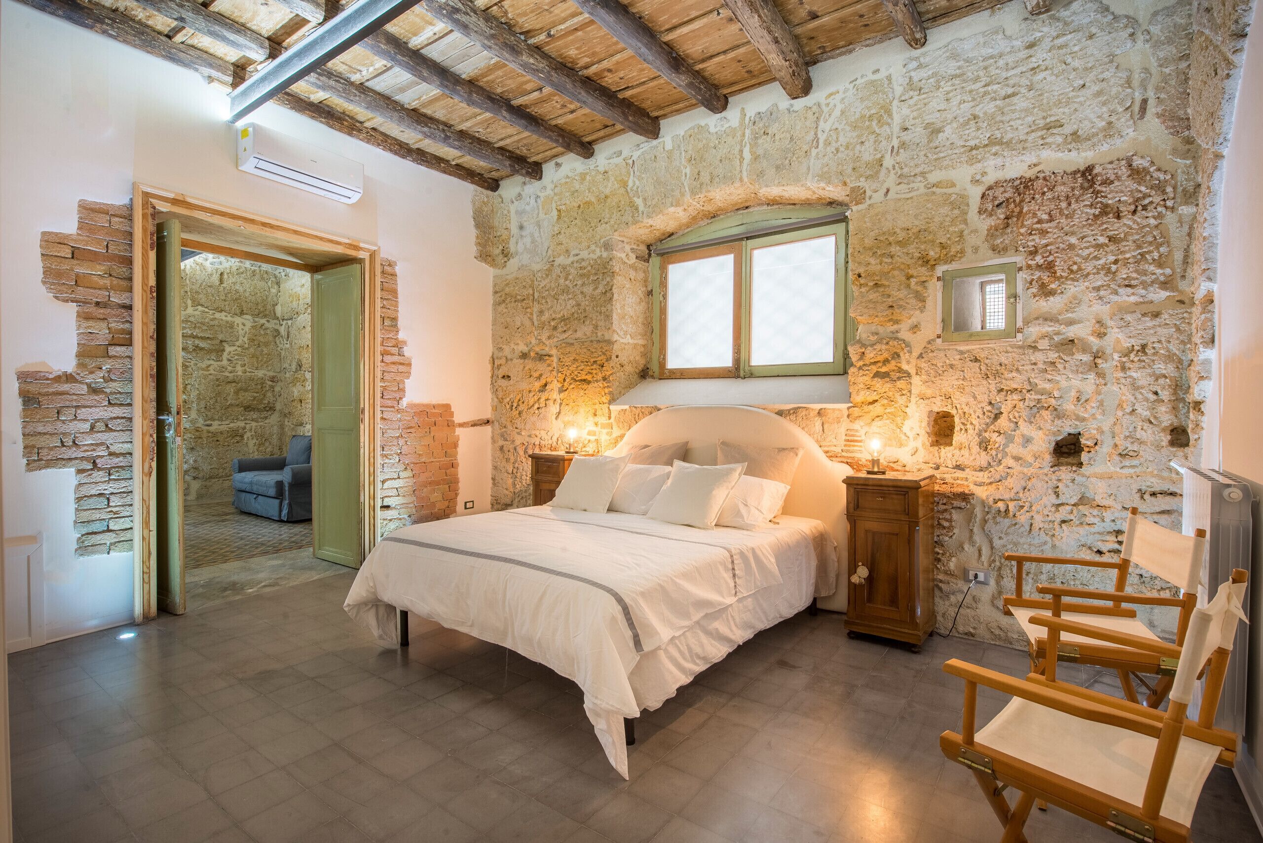 Un dormitorio con pared de piedra, una cama, una mesita de noche de madera, una silla y una ventana.