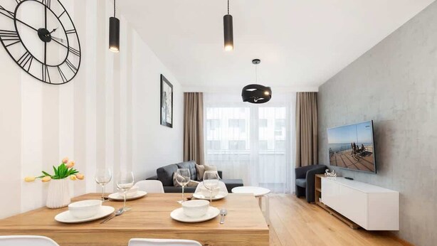 A modern dining area with a wooden table set for four, a large wall clock, and a television on the right.