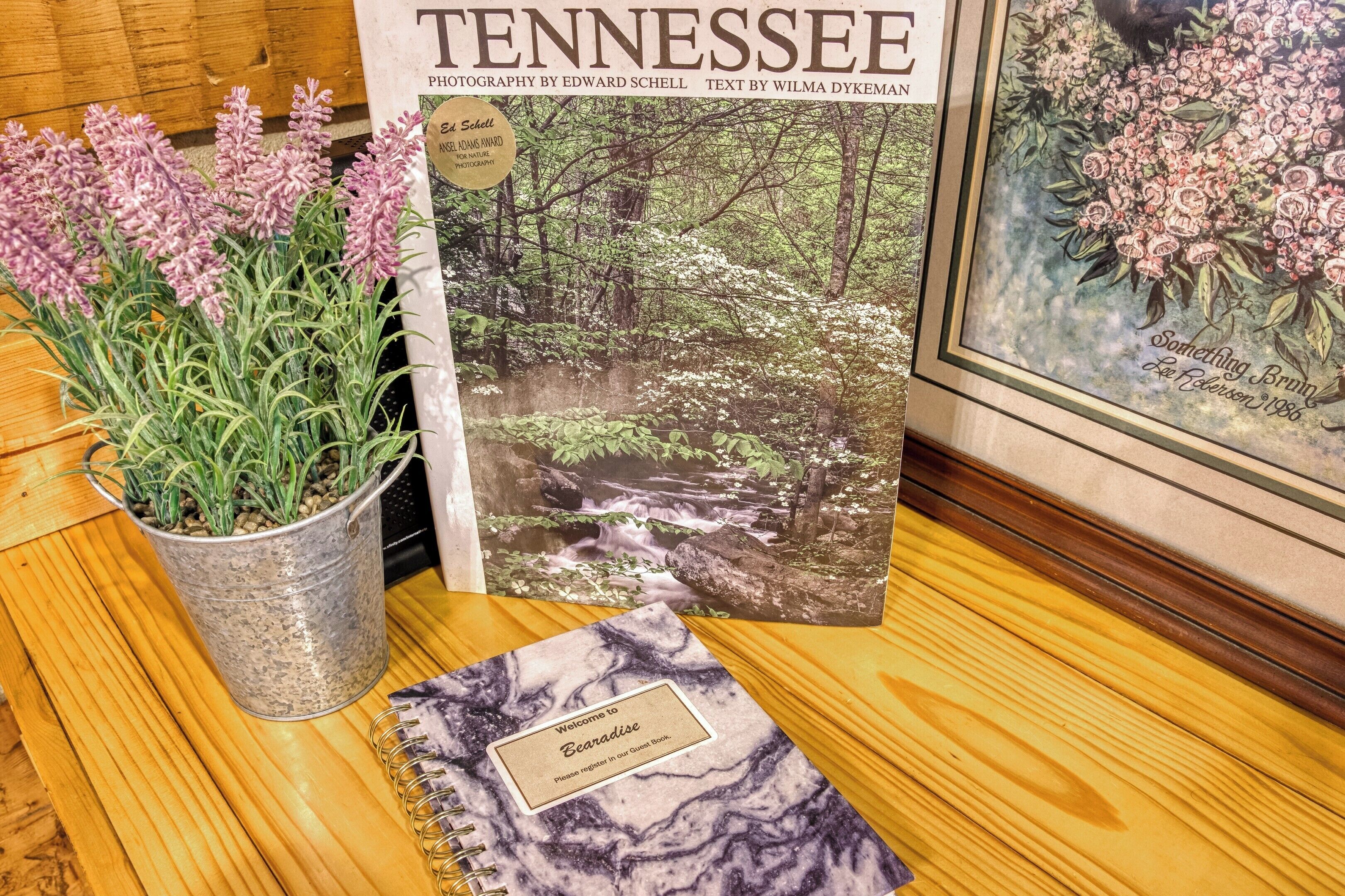 A Tennessee-themed book, a framed picture of a forest stream, and a potted plant on a wooden table.