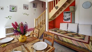 Une pièce chaleureuse avec un escalier en bois, un lit recouvert d’une courtepointe aux couleurs vives, une table à manger ornée d’un vase de fleurs et une horloge murale.