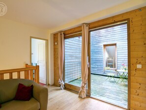 Quarto com piso de madeira, sofá bege e porta de vidro deslizante que dá para uma área externa com mesa e cadeiras.