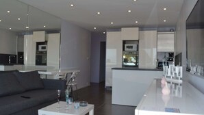 A modern living room with a grey sofa, a white coffee table, and a kitchen area with white cabinets and appliances.