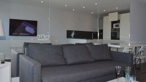 A modern living room with a grey sofa, a flat-screen TV mounted on the wall, and a kitchen area visible through a glass partition.
