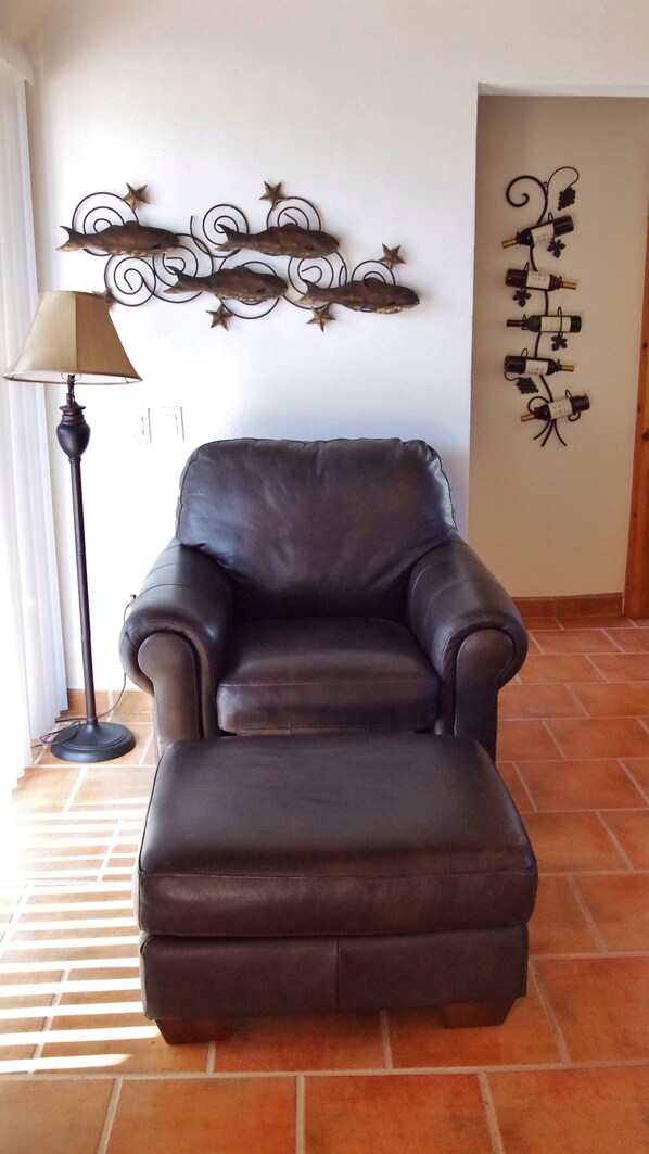Een donkerbruine leren fauteuil in een kamer met betegelde vloer, een lamp en decoratieve wandkunst.