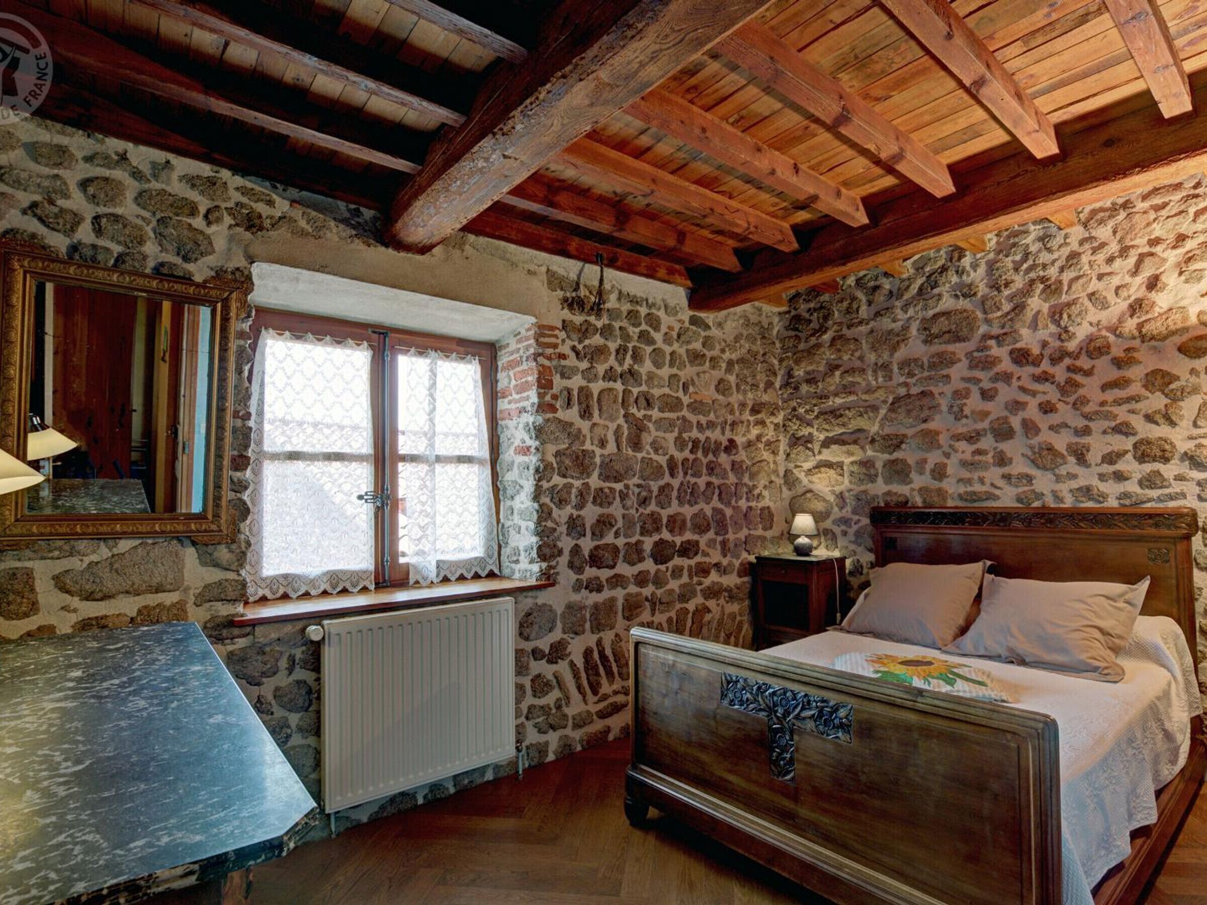 A rustic bedroom with a stone wall, wooden ceiling beams, a bed with a floral headboard, a window with lace curtains, and a small table with a lamp.