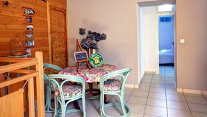A dining area with a round table and four chairs, a wooden wall with a clock, and a door leading to another room. - Gîte Bellevue in Marols for 6 people with charming enclosed yard (Marols)