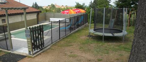 Un espacio al aire libre cercado con piscina, trampolín y zona de estar cubierta.