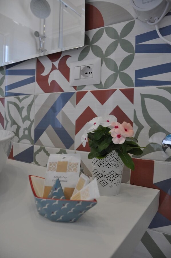 Een badkamer met een betegelde spatwand, een witte wastafel, een witte vaas met bloemen en een kom met doucheproducten.