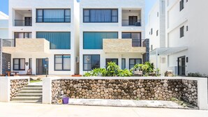 A modern multi-story residential building with balconies, large windows, and a stone wall with logs in front. - Penghu Water Reflection (Magong)