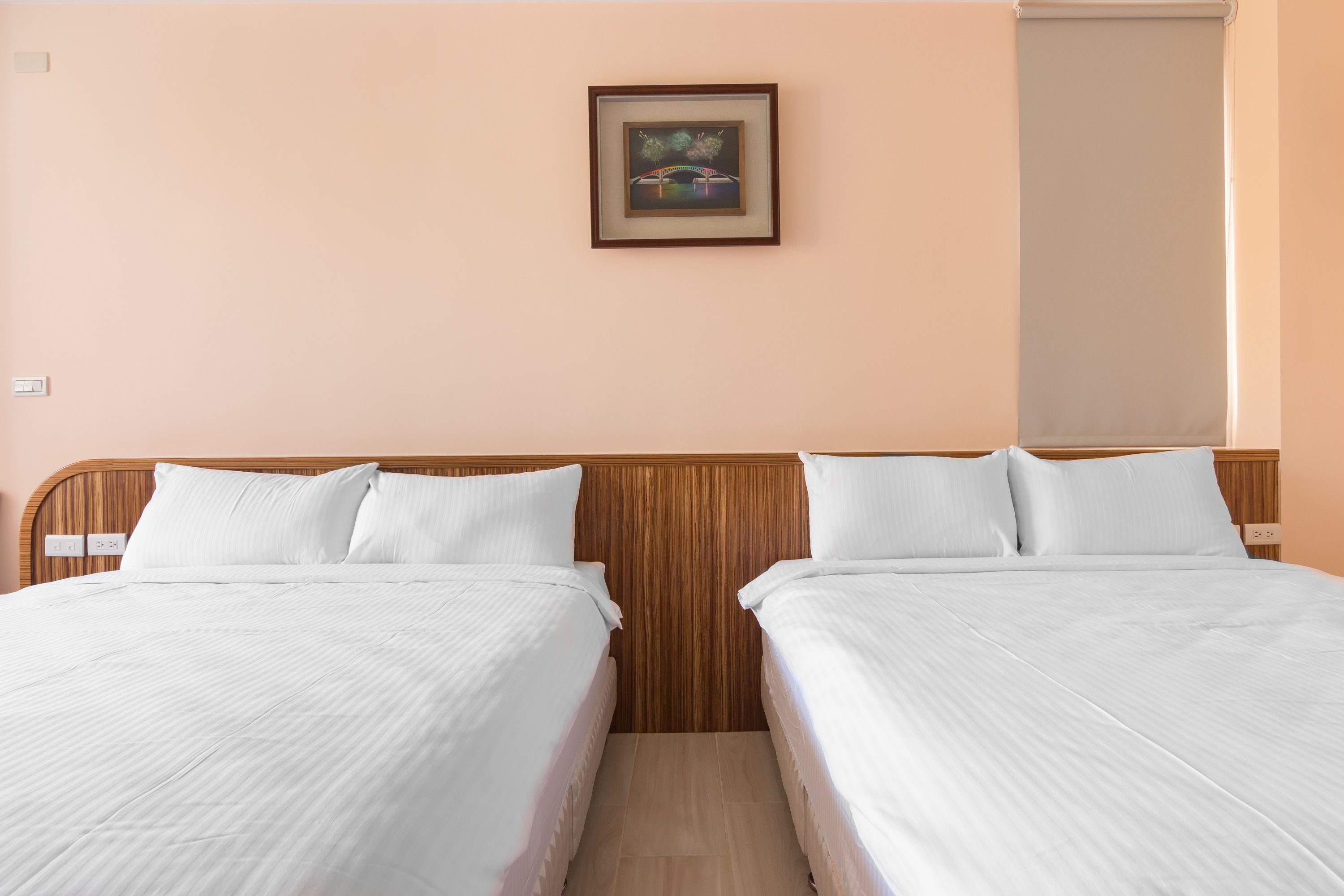 Een hotelkamer met twee bedden, een houten hoofdbord, een lijst met een foto aan de muur en een raam met een zonwering.