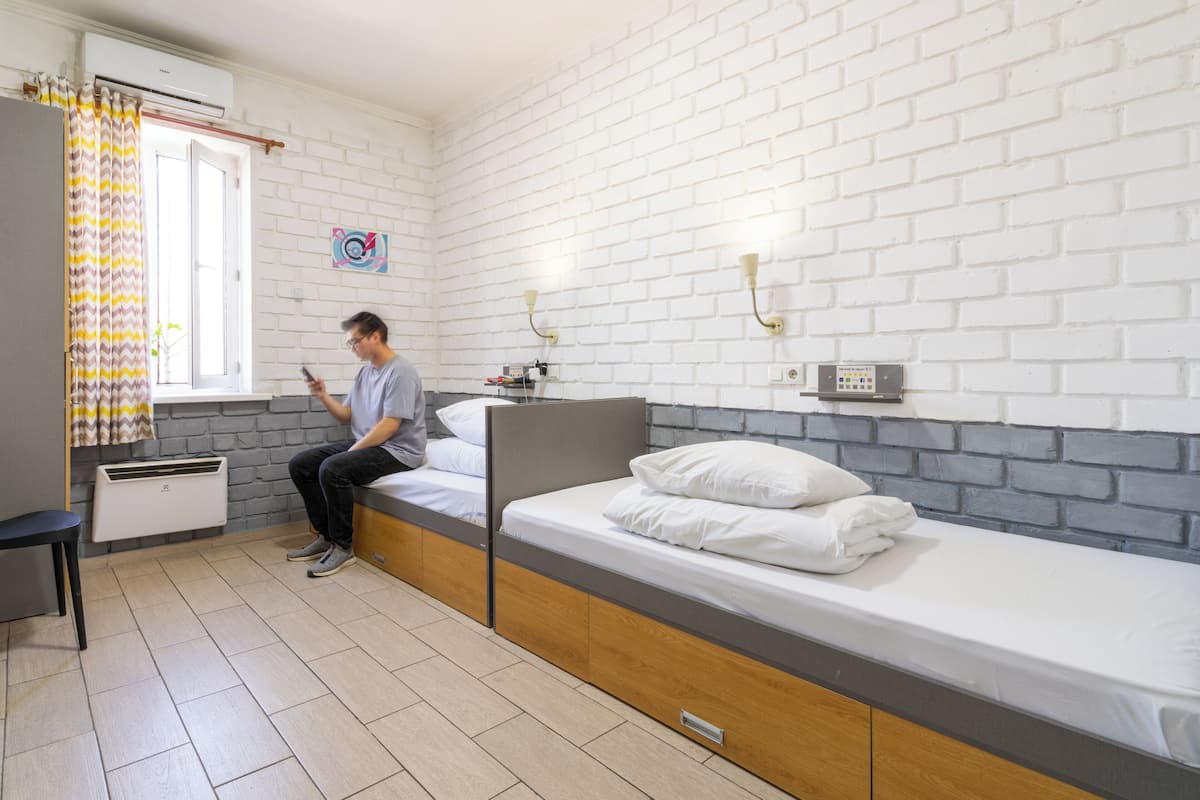 A single-bed room with a white brick wall, a window with curtains, a bedside table, and a wall-mounted lamp.