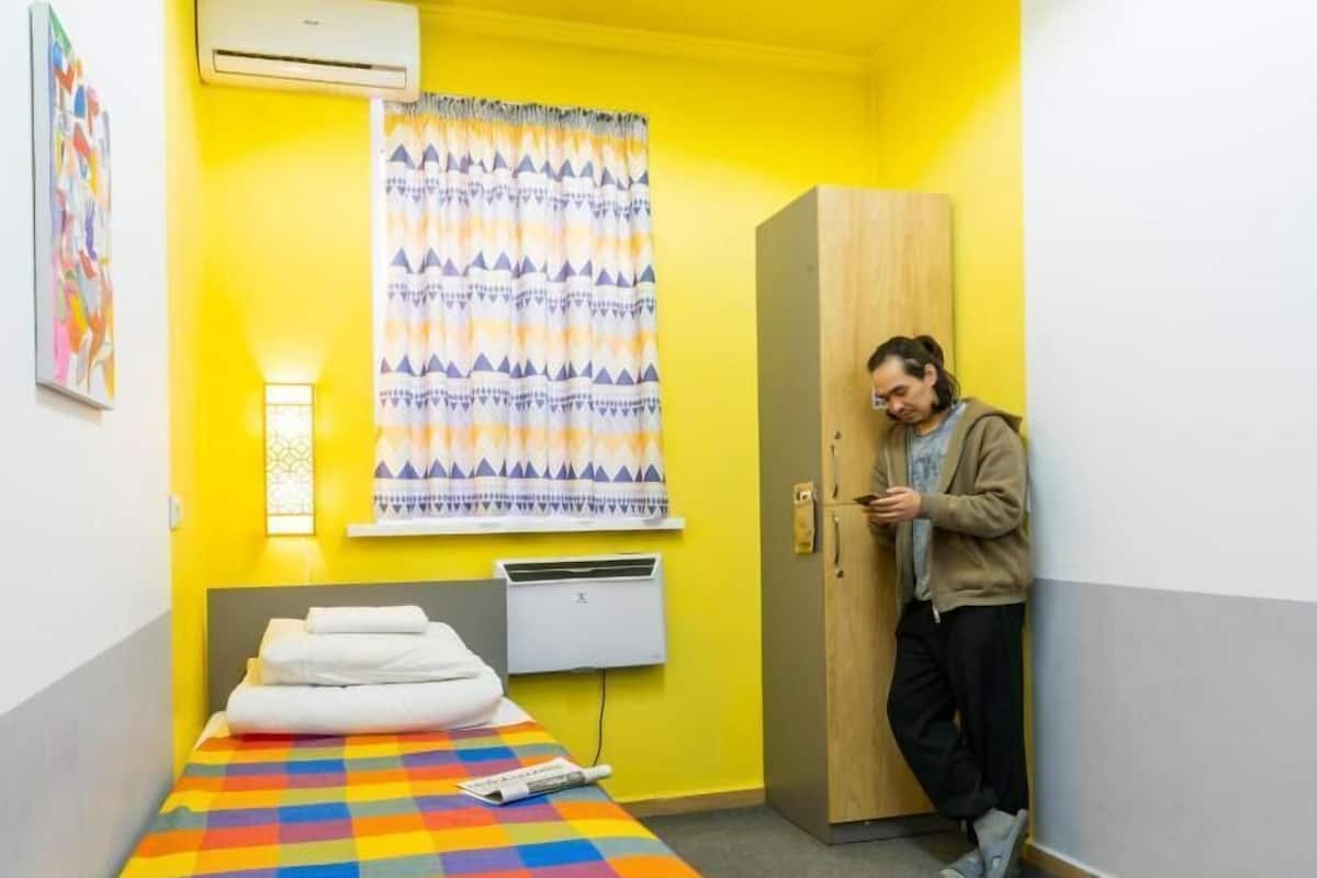 A small room with a bed, a wardrobe, an air conditioning unit, and a window with curtains.